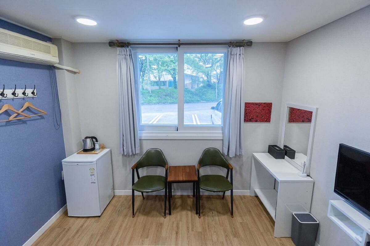 Een compacte hotelkamer met een klein tafeltje en twee stoelen, een minibar, een televisie, een raam met gordijnen en een kapstok met hangers.