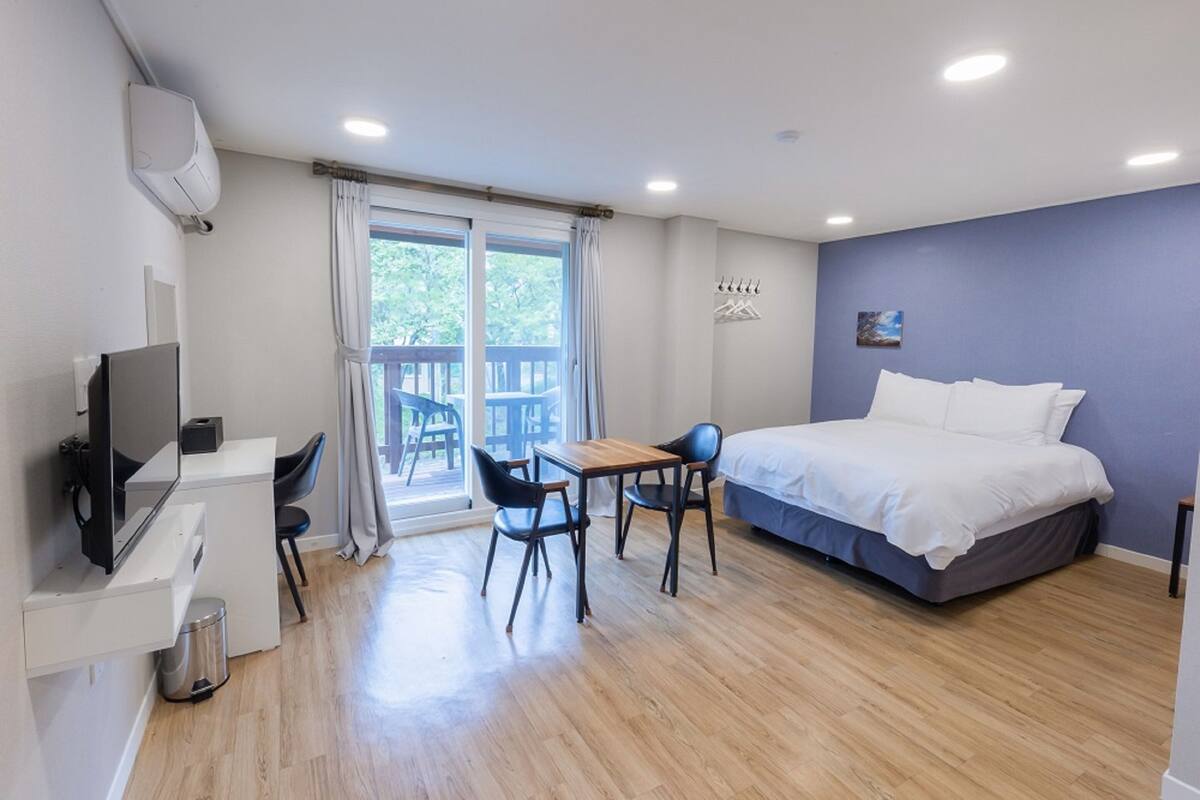 Een moderne hotelkamer met een bed, een bureau met stoel, een televisie en een balkon met uitzicht op bomen.
