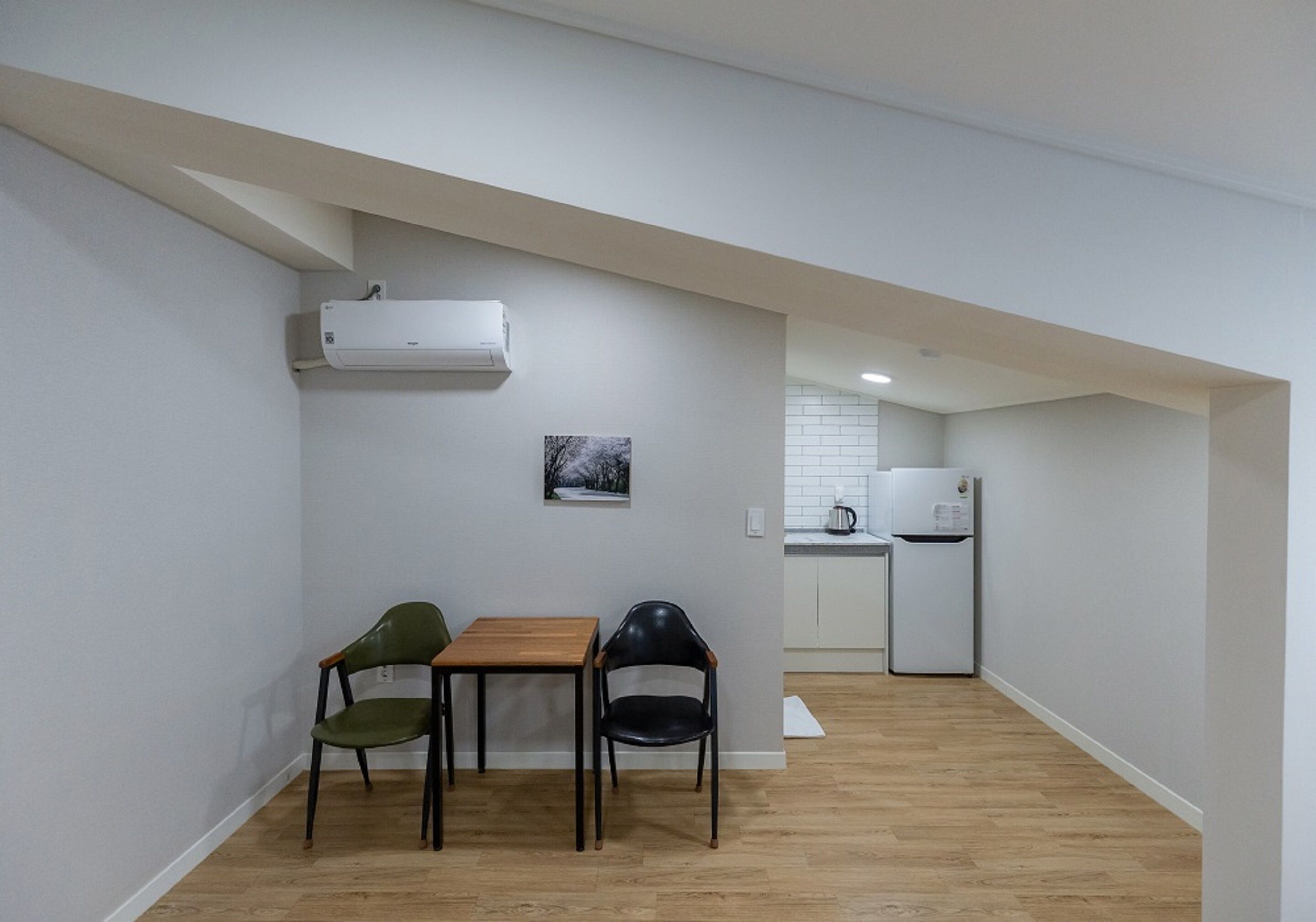 A small, open-plan living and dining area with a wooden table and two chairs, a kitchen in the background, and a white air conditioning unit on the wall.