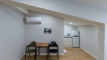 A small, open-plan living and dining area with a wooden table and two chairs, a kitchen in the background, and a white air conditioning unit on the wall.