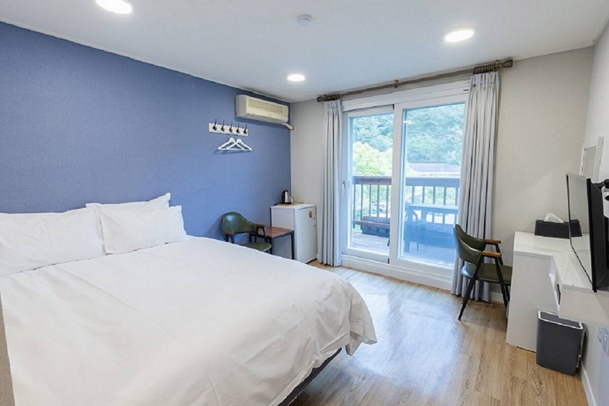 Hotelkamer met een groot bed, een bureau, een stoel en een kleine tafel. Er is een raam met gordijnen en uitzicht op groen.