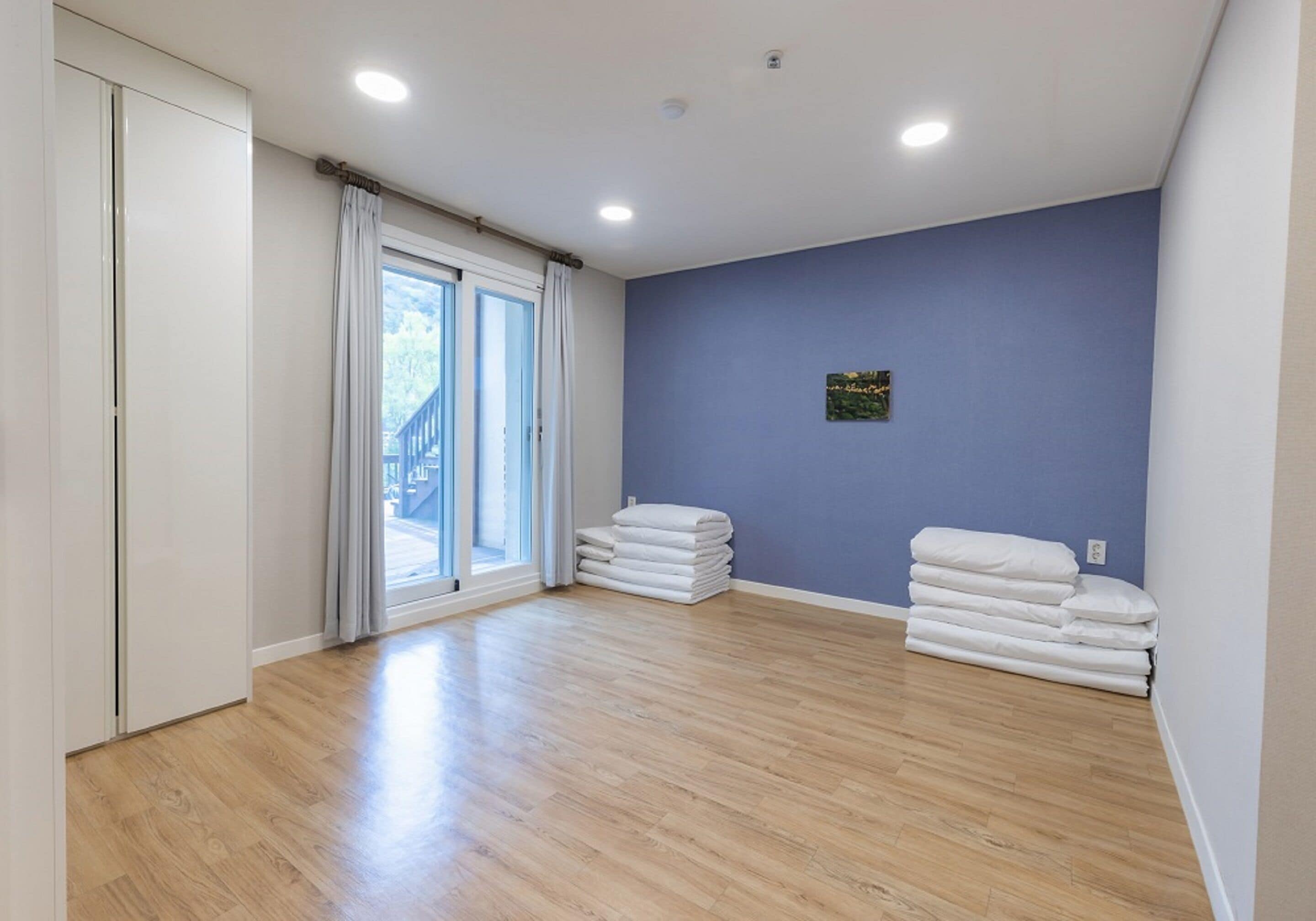 A room with wooden flooring, a large window with curtains, a blue wall, and a small picture.