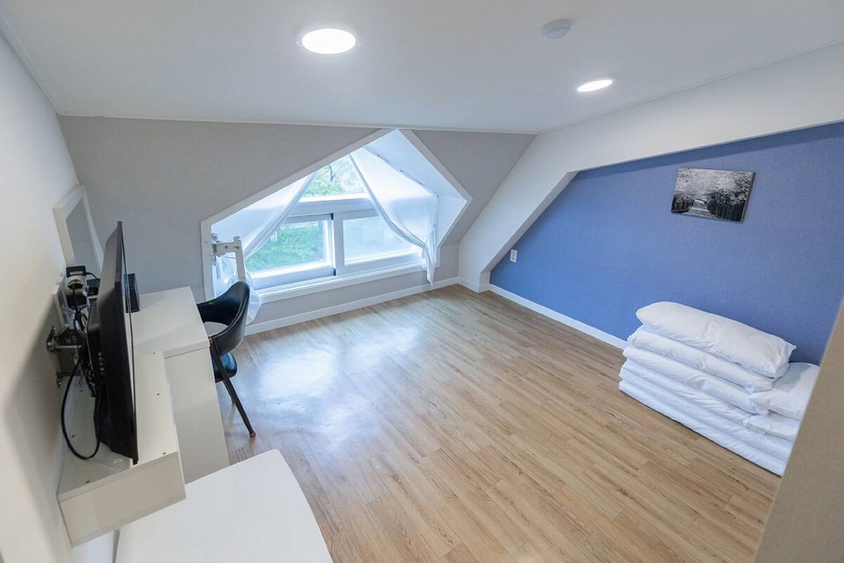 Een kamer met een schuine plafond, een groot raam, een bureau met stoel, en een stapel kussens tegen een blauwe muur.