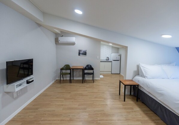 Habitación de hotel moderna con televisión, un pequeño comedor con sillas, piso de madera, cama y una kitchenette al fondo.