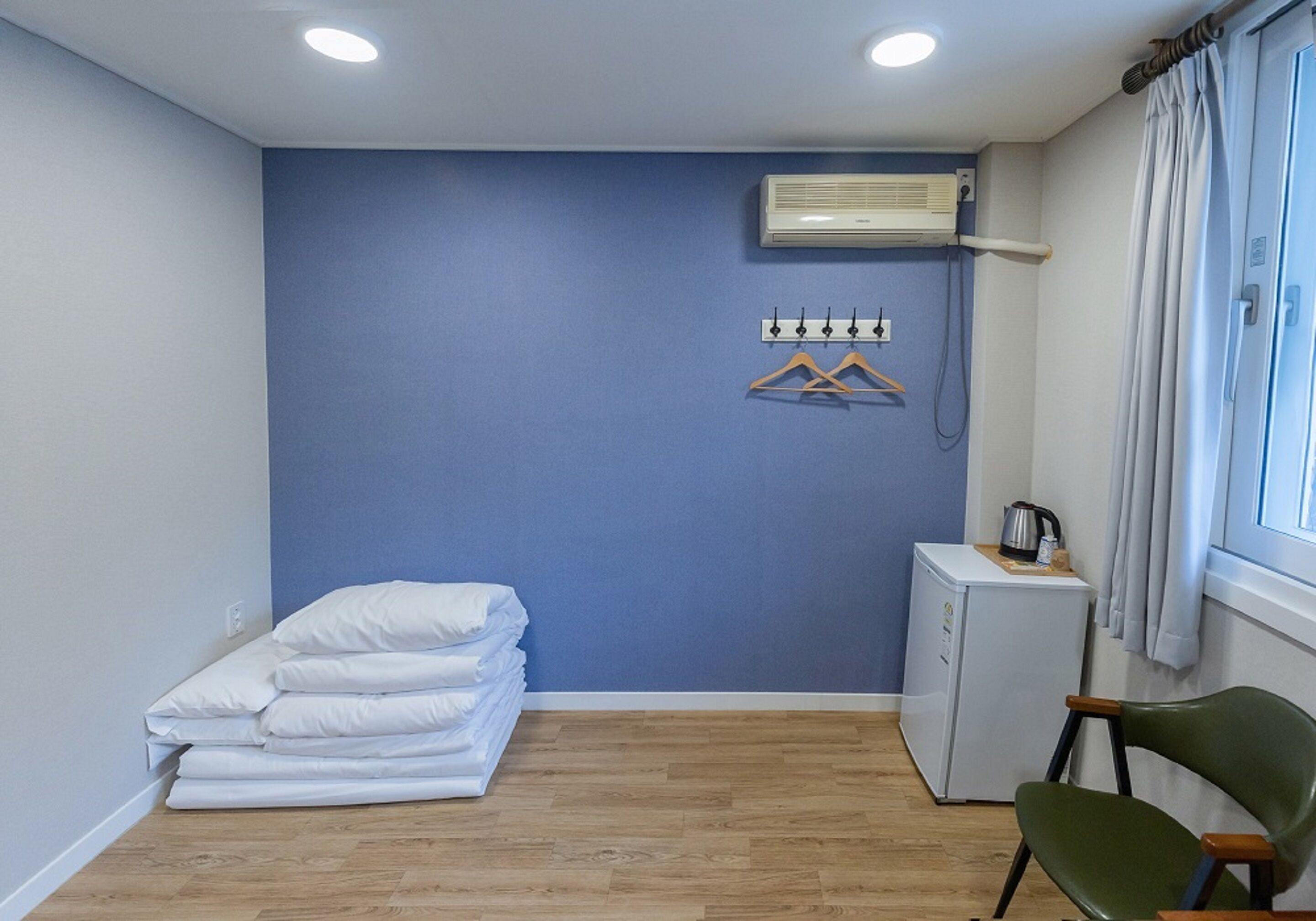 A room with a blue wall, a bed with white linens, a small kitchenette, and a chair.
