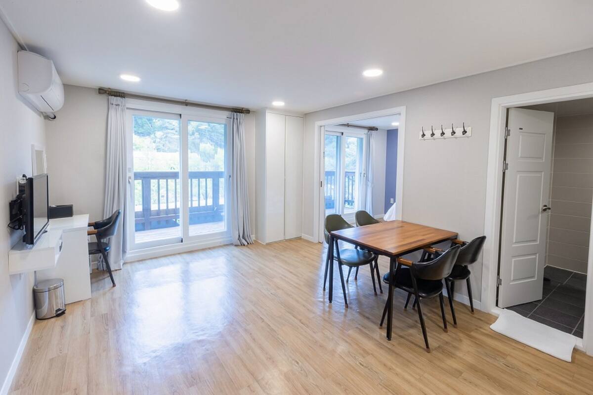 Een ruime kamer met een houten eettafel, stoelen, een televisie en een balkon met gordijnen.