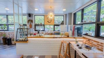 Interior moderno de una cafetería con mostrador, zona de estar y grandes ventanales que ofrecen vistas al verdor.
