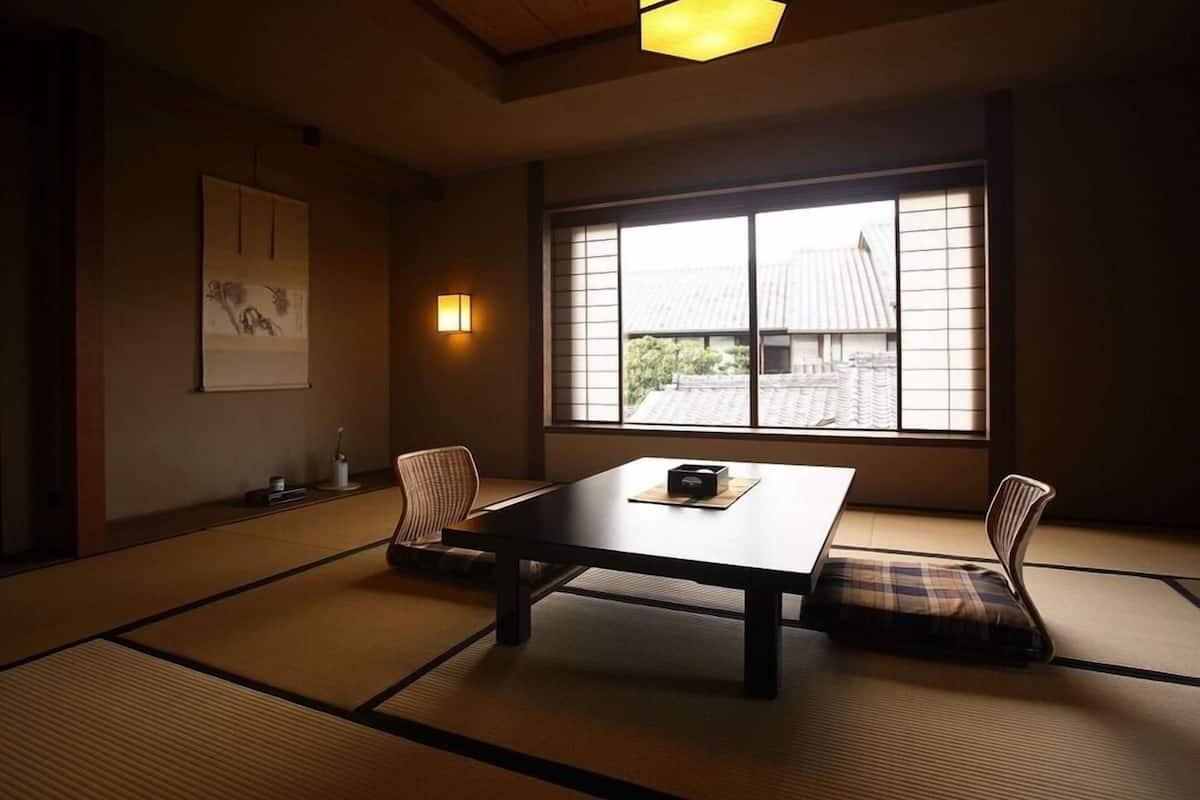 A traditional Japanese room with tatami flooring, a low table, two chairs, a hanging light fixture, and a window with a view of traditional buildings.