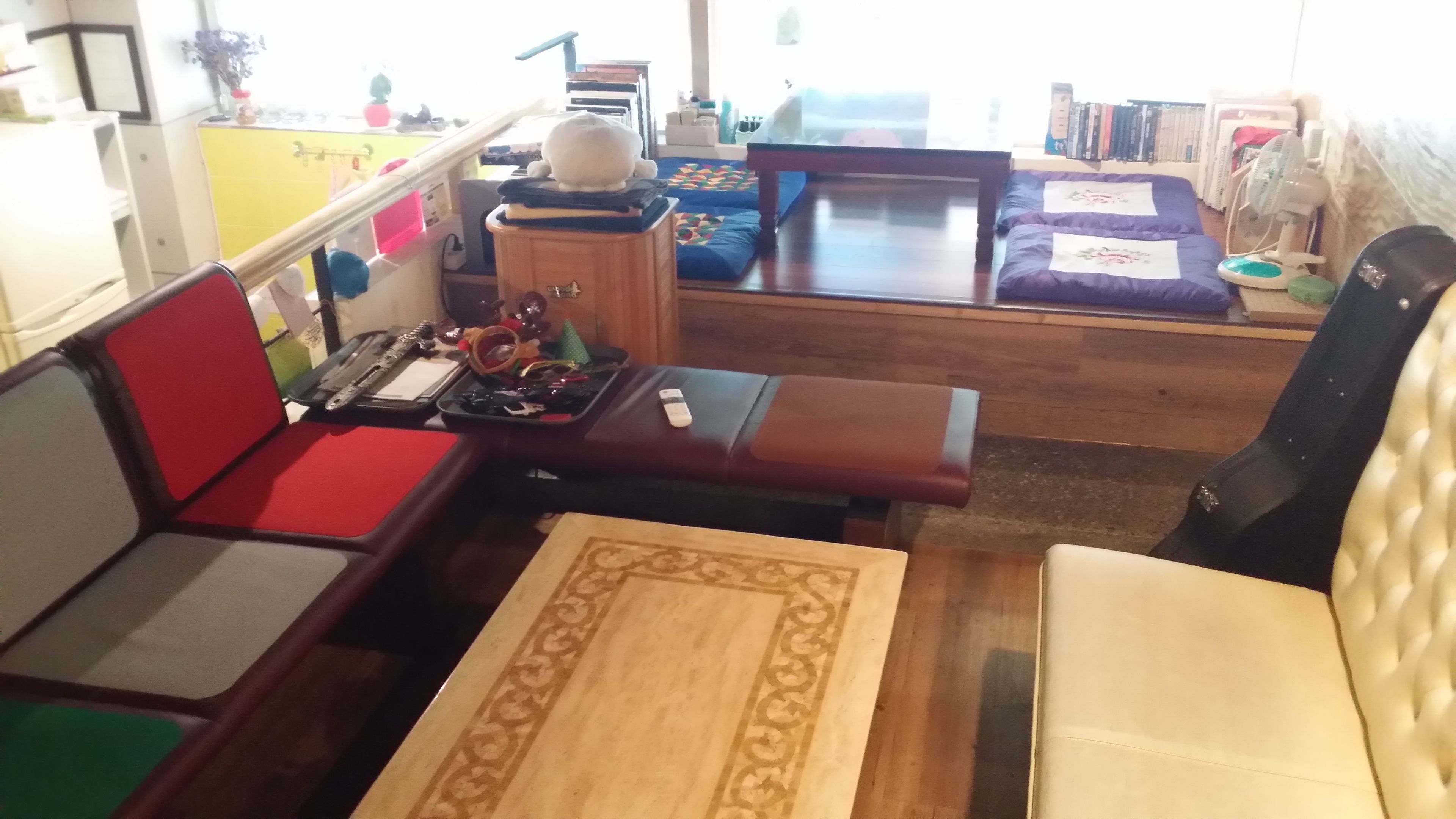 A compact living space with a sofa, a small table, and a bench. There is a window providing natural light, and a bookshelf filled with books.