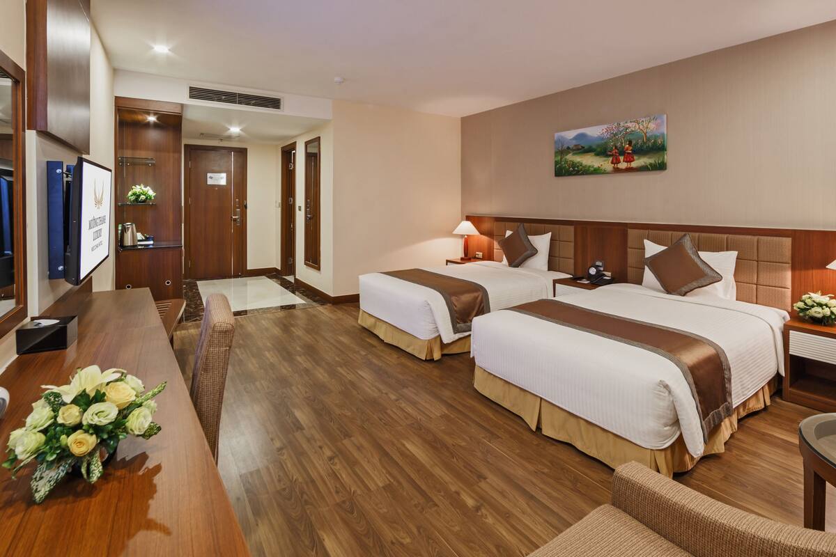 A hotel room with two beds, a wooden desk, a chair, a TV, and a painting on the wall.