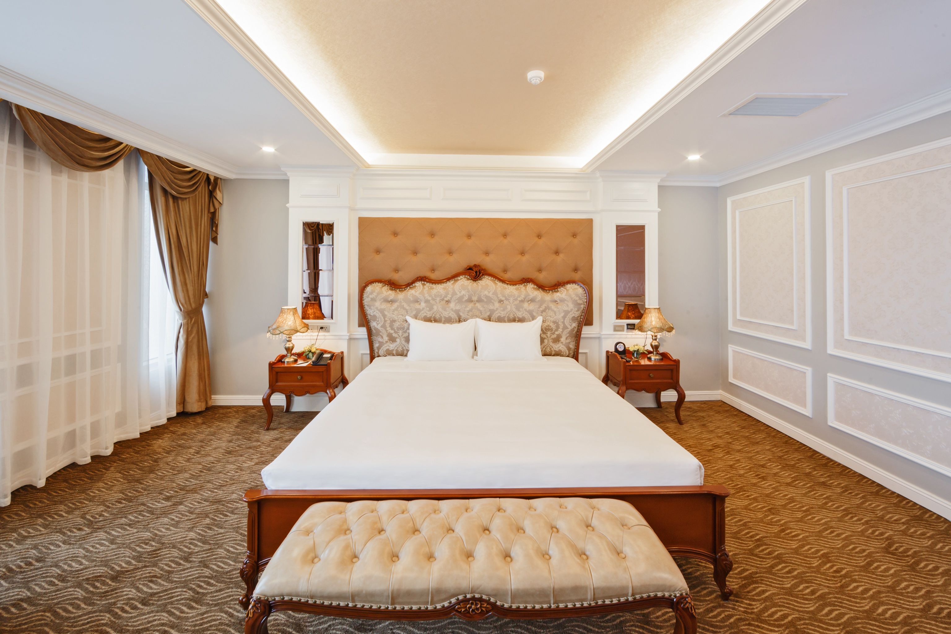 A large, centrally positioned bed with a tufted headboard, flanked by two bedside tables with lamps, in a room with patterned carpet and a ceiling light fixture.