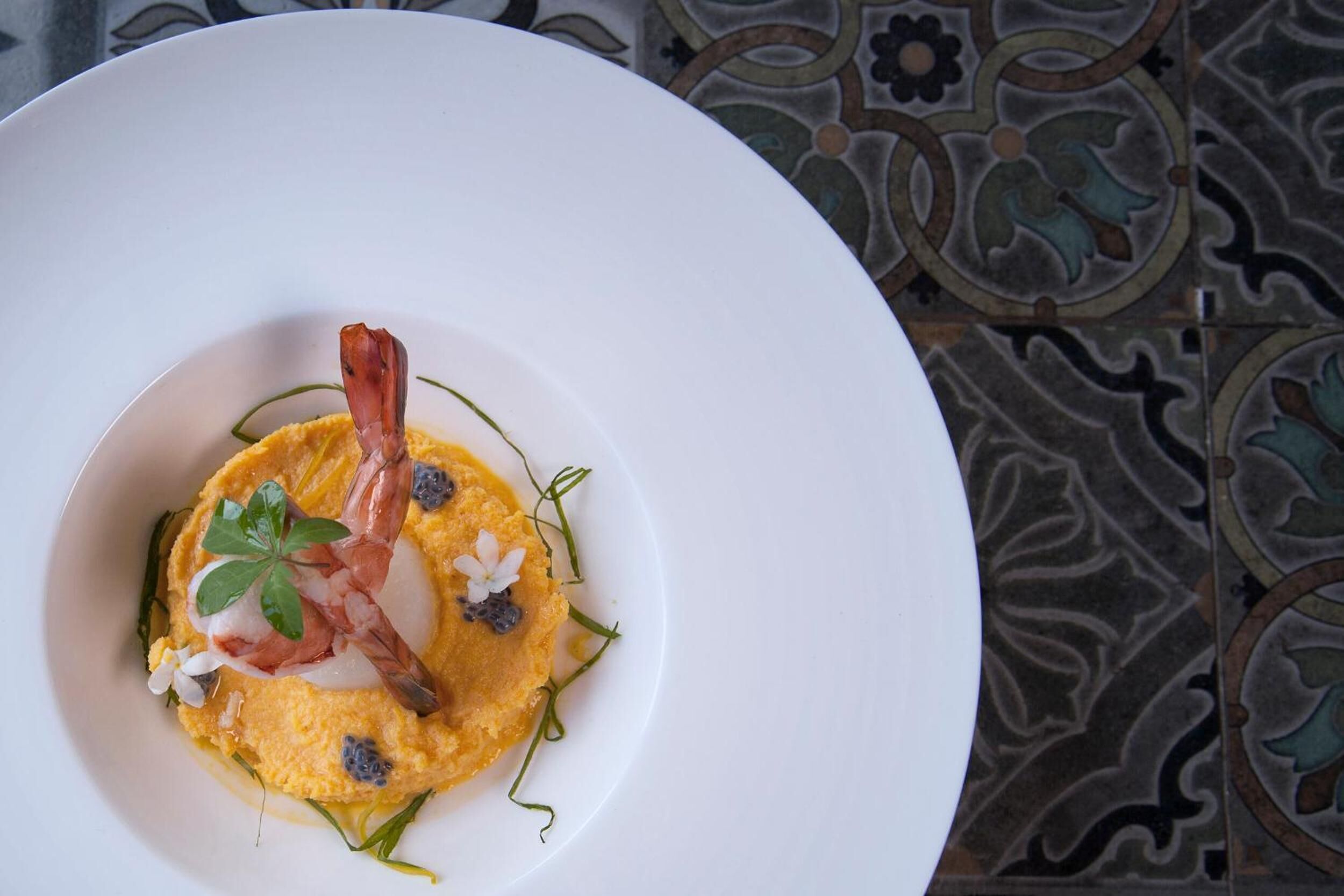 A white plate with a yellow dish, garnished with a piece of bacon, white flowers, and green leaves.