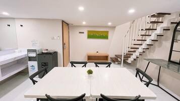 A modern dining area with a white table, black chairs, a small plant, a water dispenser, and a staircase.