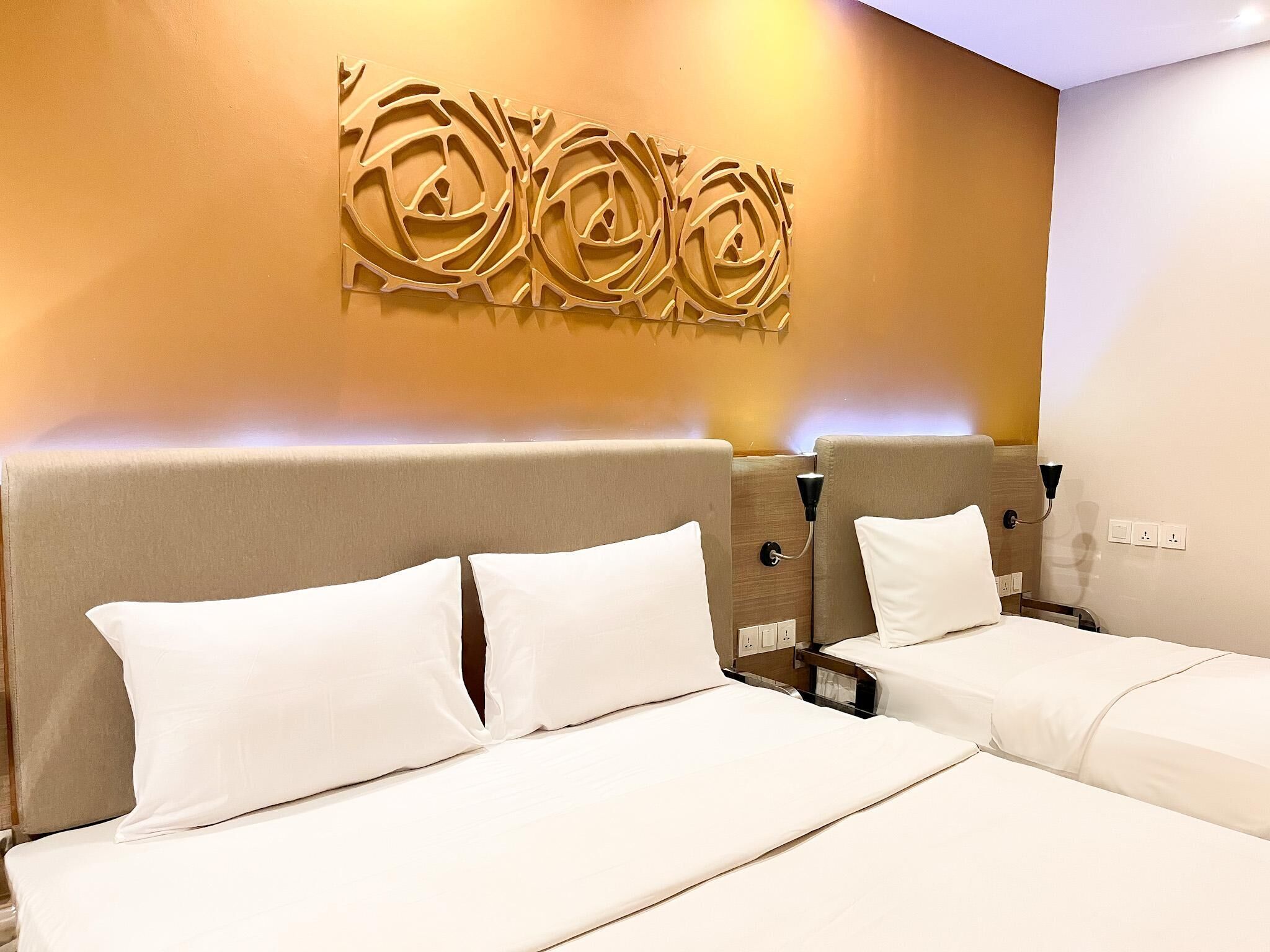 A hotel room with two beds, a decorative wall panel, and a bedside table with a lamp.