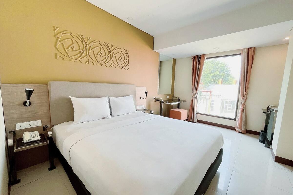 Ein Hotelzimmer mit einem großen Bett, einem Nachttisch mit Lampe, einem kleinen Tisch mit Spiegel und einem Fenster mit Vorhängen.