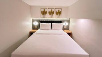 Une chambre d’hôtel avec un grand lit, deux tables de chevet, une tête de lit recouverte d’un tissu foncé et trois appliques murales.