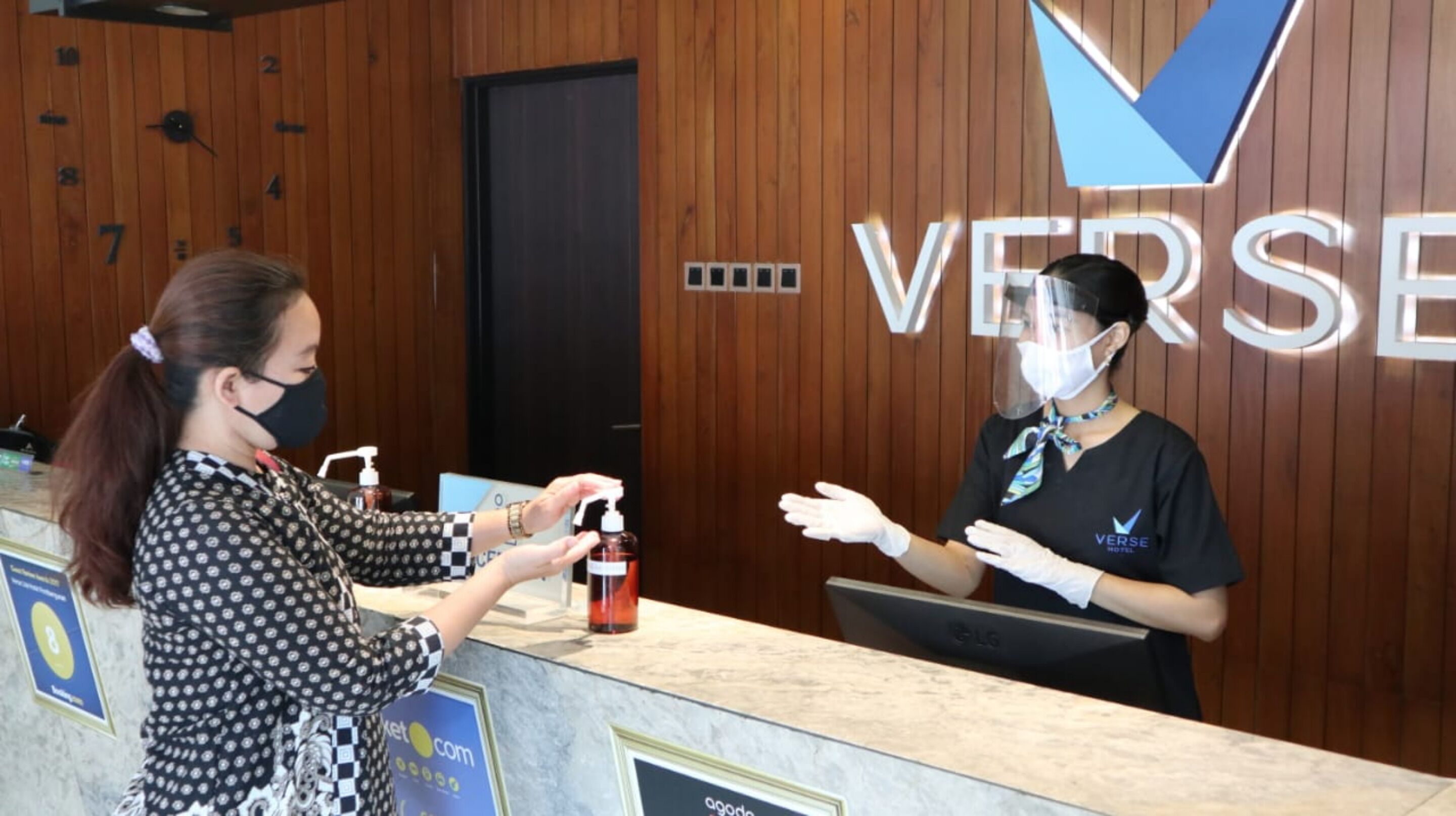 A hotel reception area with wooden paneling and a reception desk. A guest is interacting with a staff member who is wearing a face shield and gloves.