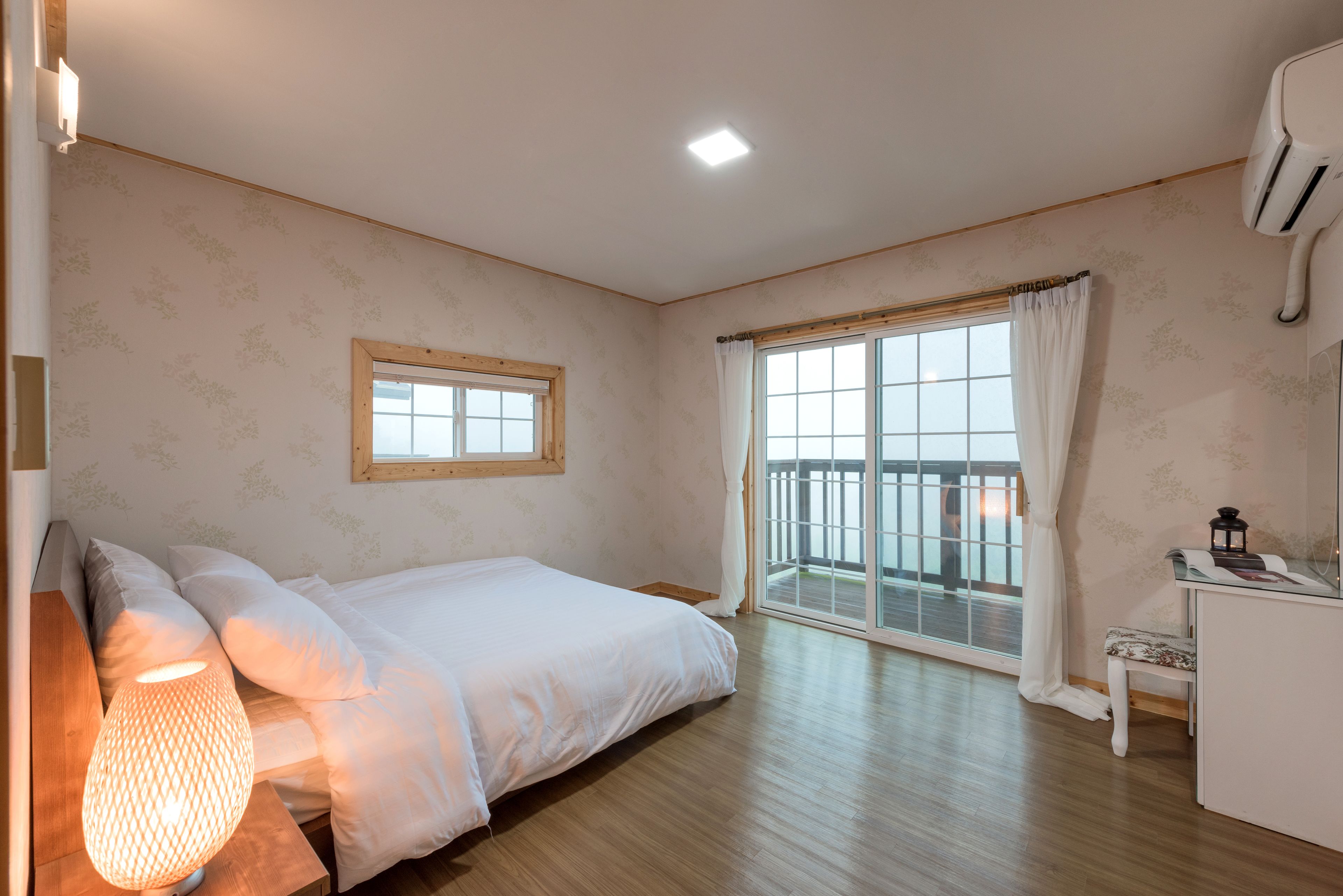 Een slaapkamer met een bed, nachtkastje, lamp, raam met gordijnen en uitzicht.