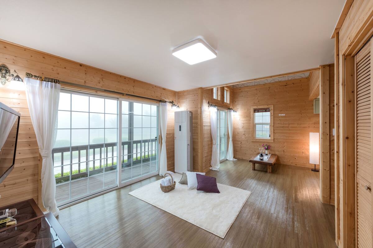 A spacious room with wooden walls and floors, large sliding glass doors to a balcony, a white rug, and a small table with a lamp.