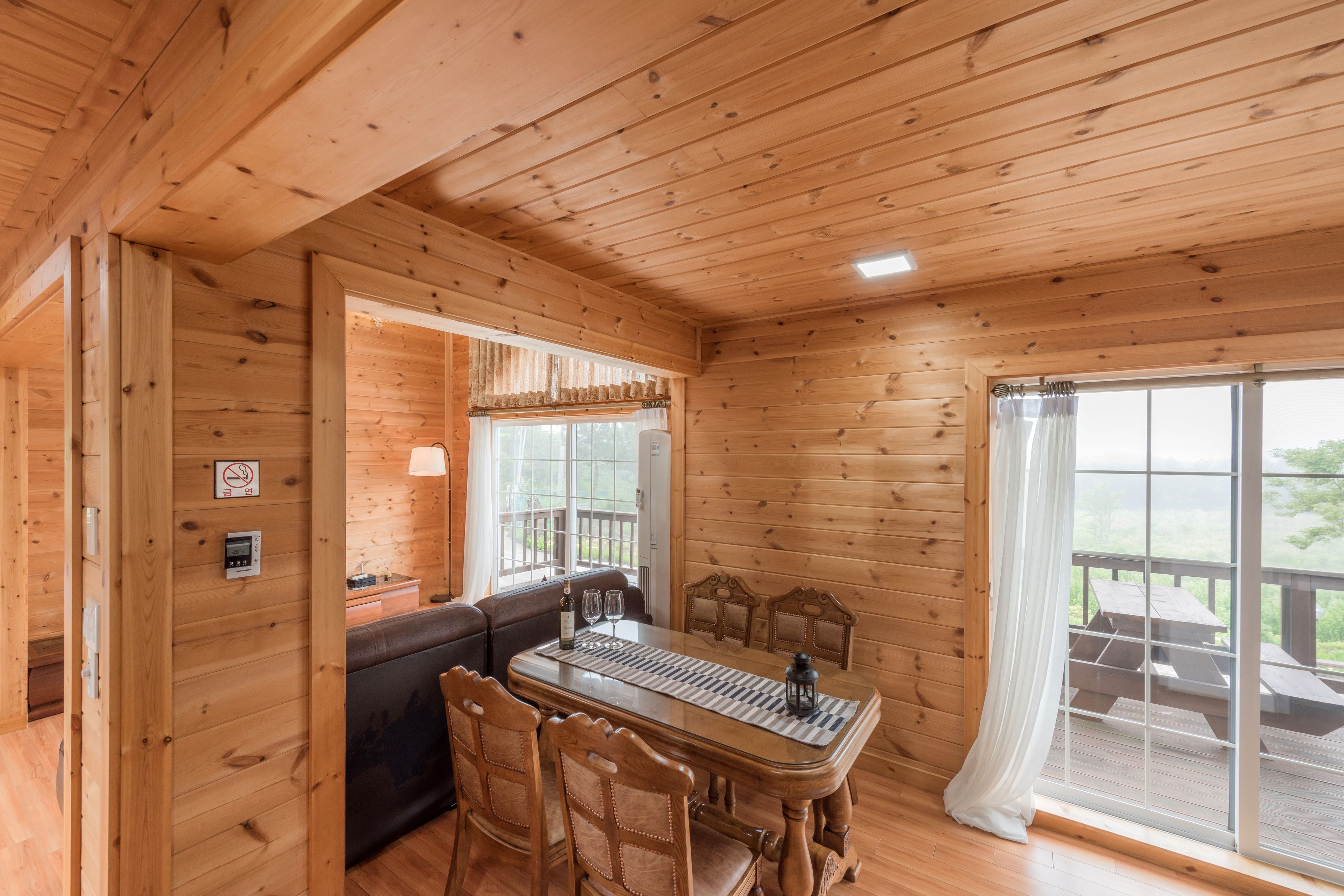 A wooden cabin interior with a dining table, chairs, a sofa, and a large window with curtains.