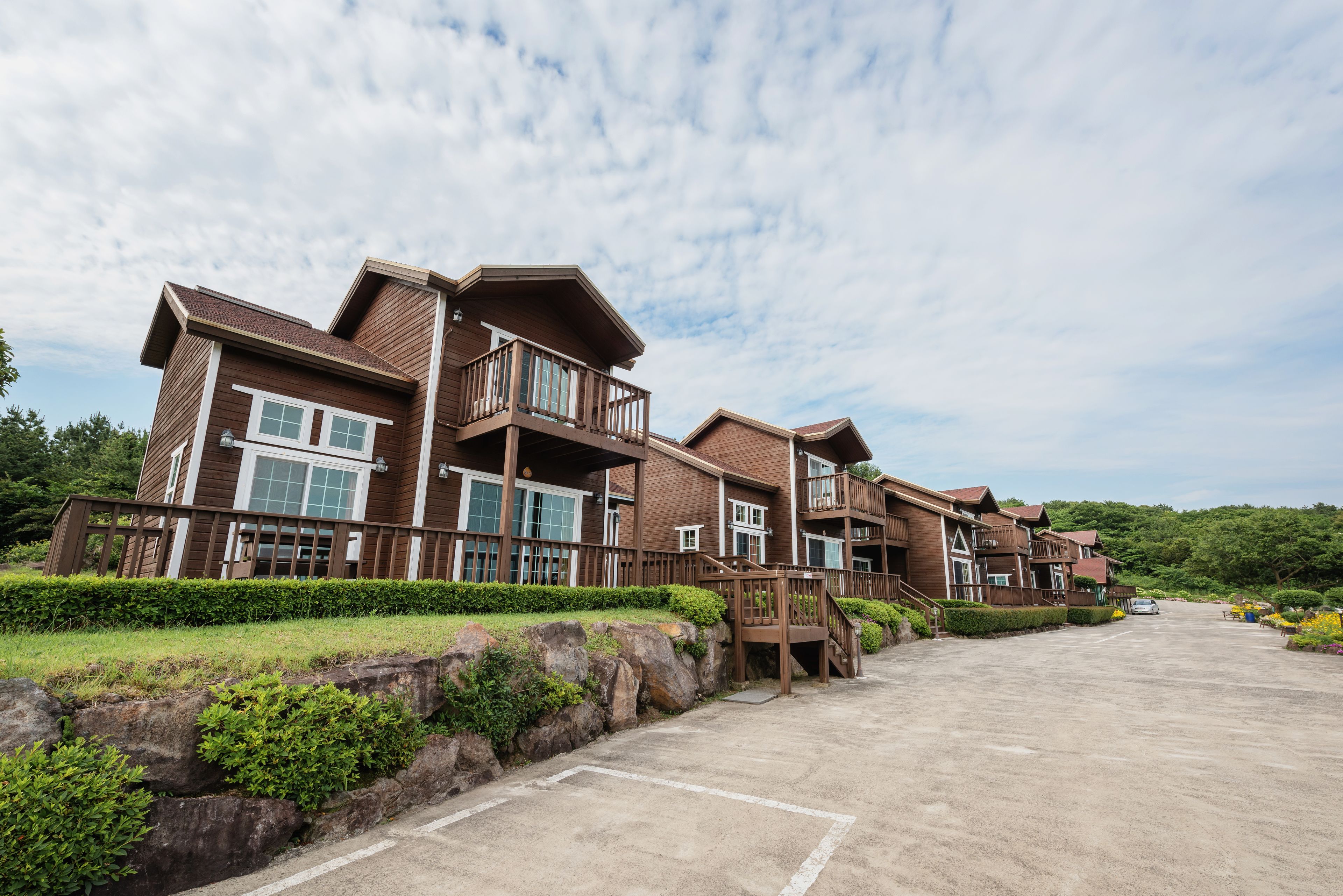 A row of multi-story wooden buildings with balconies, a parking lot, and a landscaped area with rocks and shrubs.