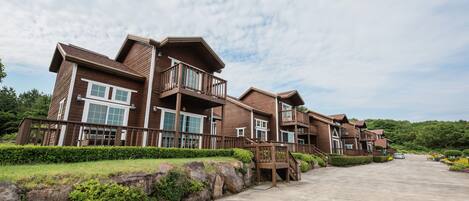 A row of multi-story wooden buildings with balconies, a parking lot, and a landscaped area with rocks and shrubs.