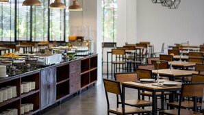 A spacious dining area with multiple tables and chairs, a long counter with food displays, and pendant lighting. - iChapter Hotel Suvarnabhumi (Bang Phli)