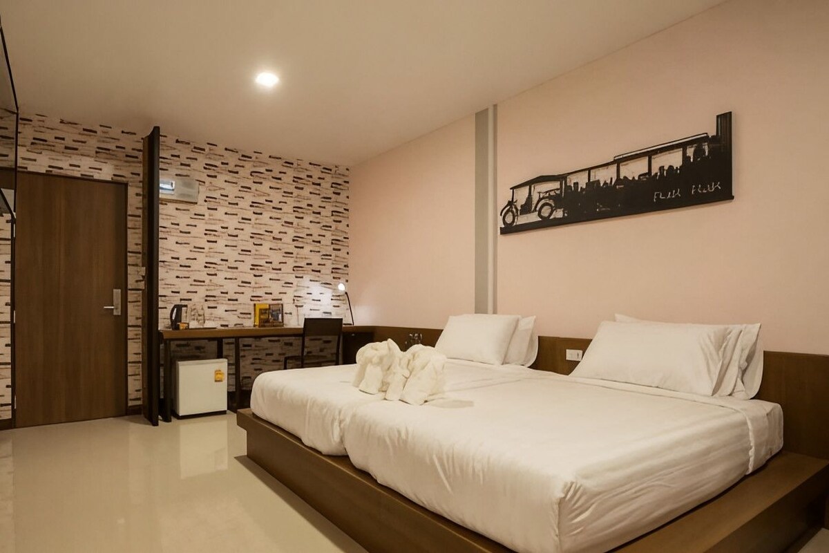 A hotel room with a bed, a desk, a refrigerator, and a wall with a cityscape artwork.
