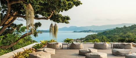 A wooden deck with beige cushions, a table, and hanging birdcages overlooking a lake and mountains.