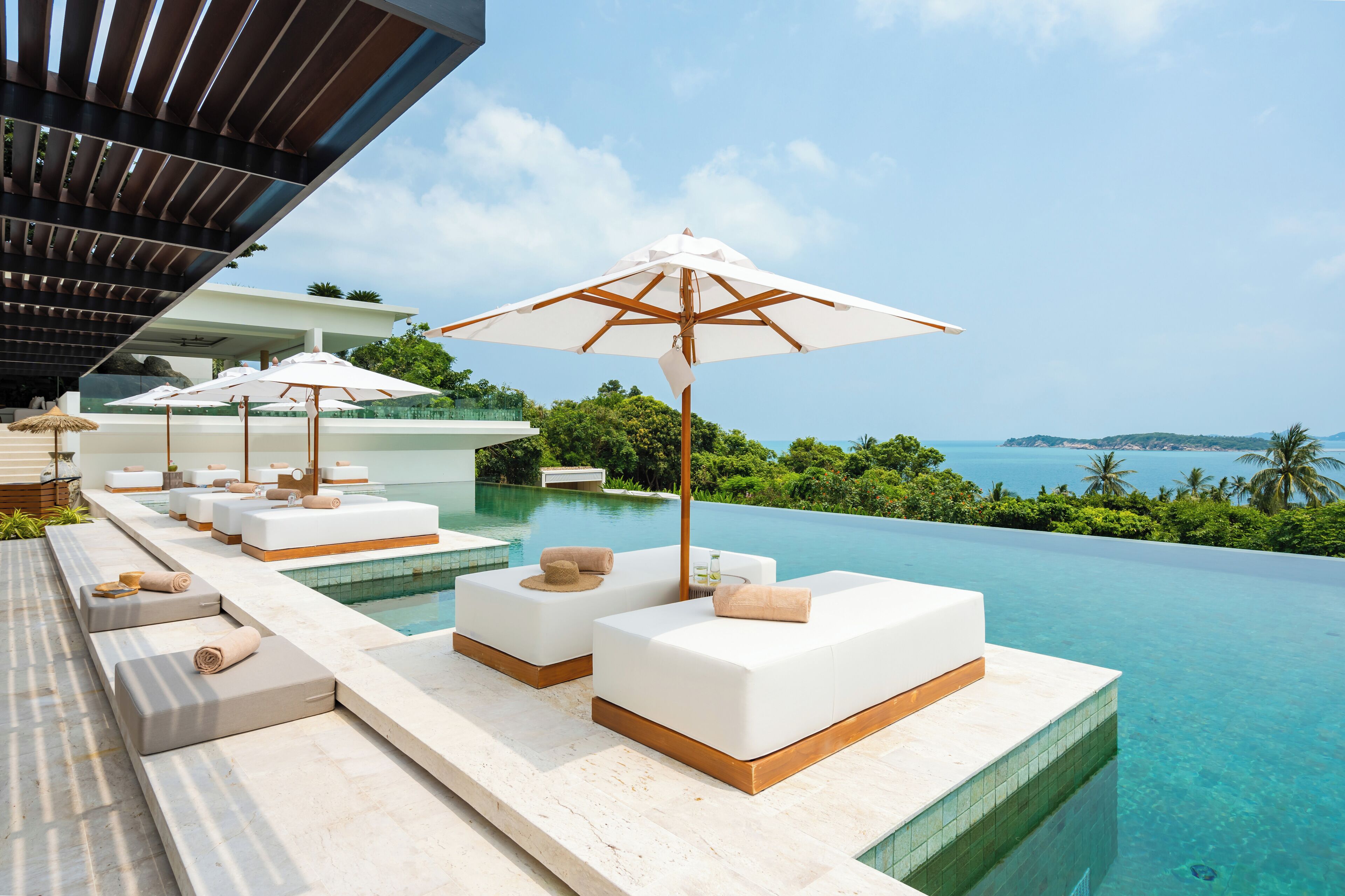 A poolside area with white lounge chairs and umbrellas, a clear view of the ocean, and a modern architectural design.