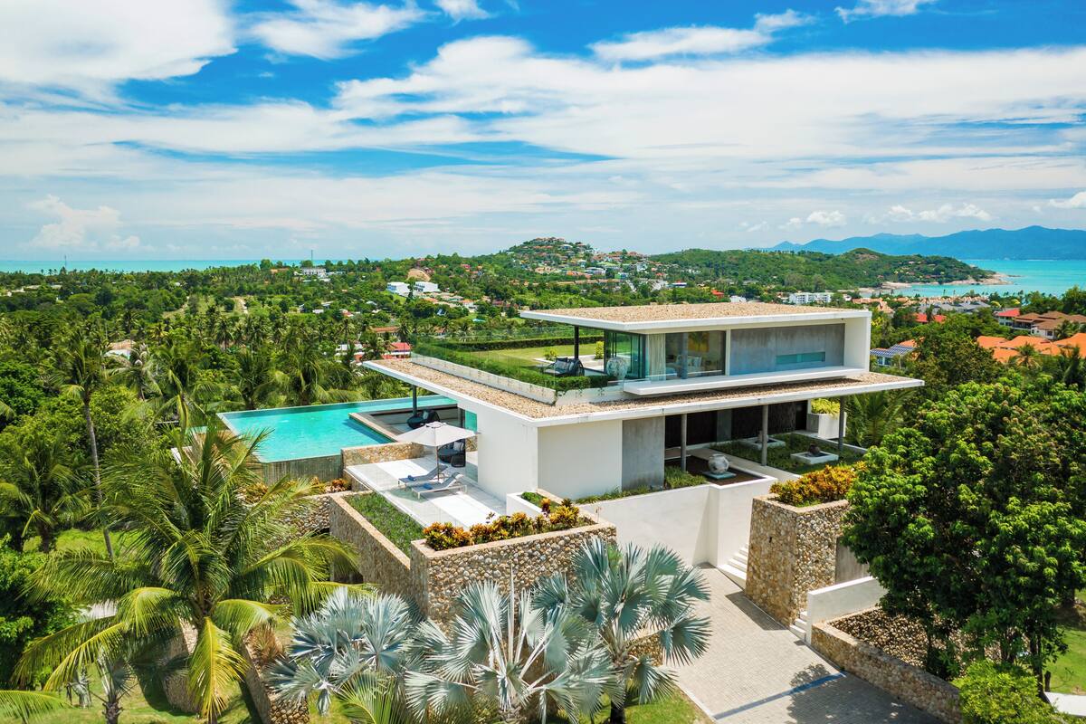 A modern villa with a rooftop pool, lush greenery, and a view of the coastline.