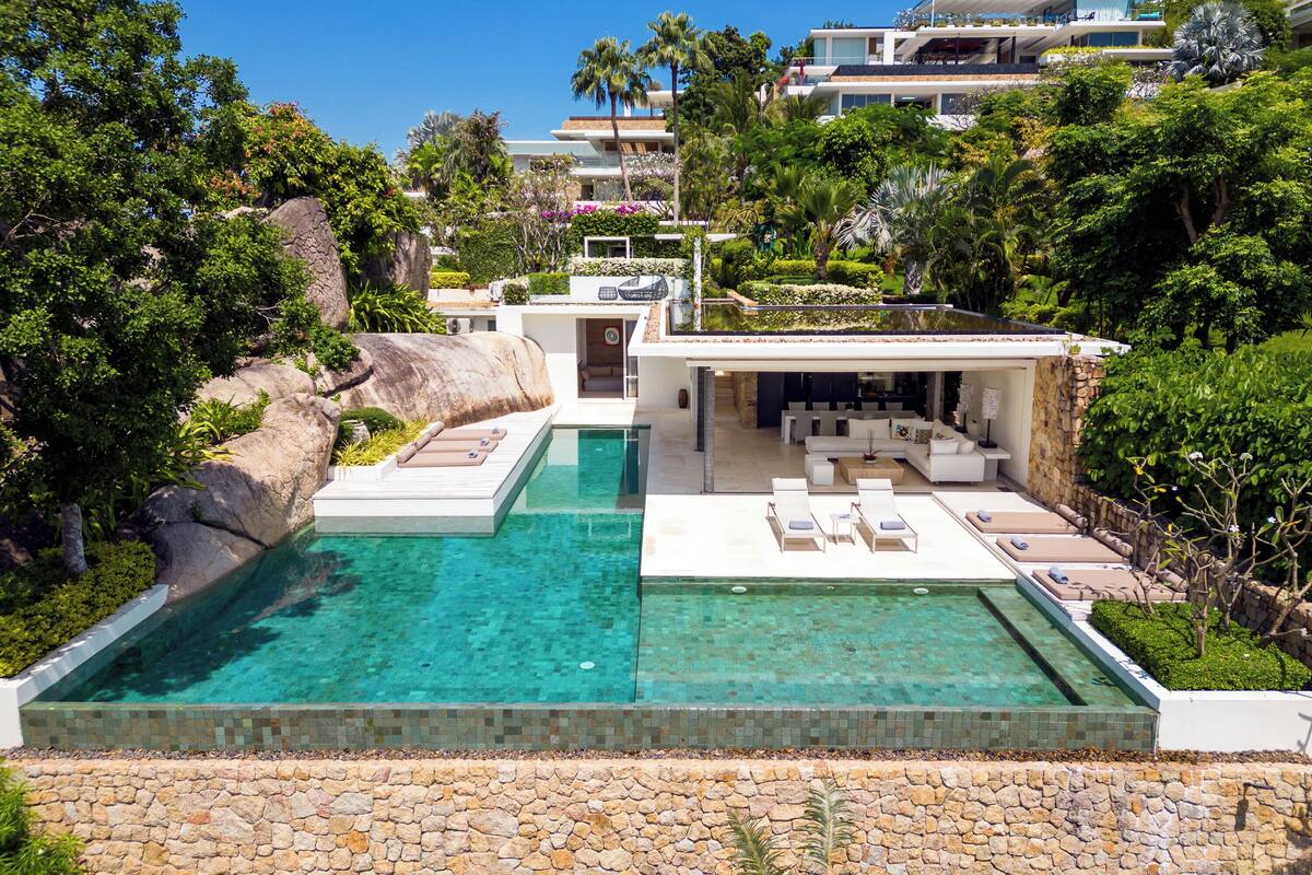 A luxurious pool area with a clear blue pool, a sun terrace with lounge chairs, and a modern building with a bar area.