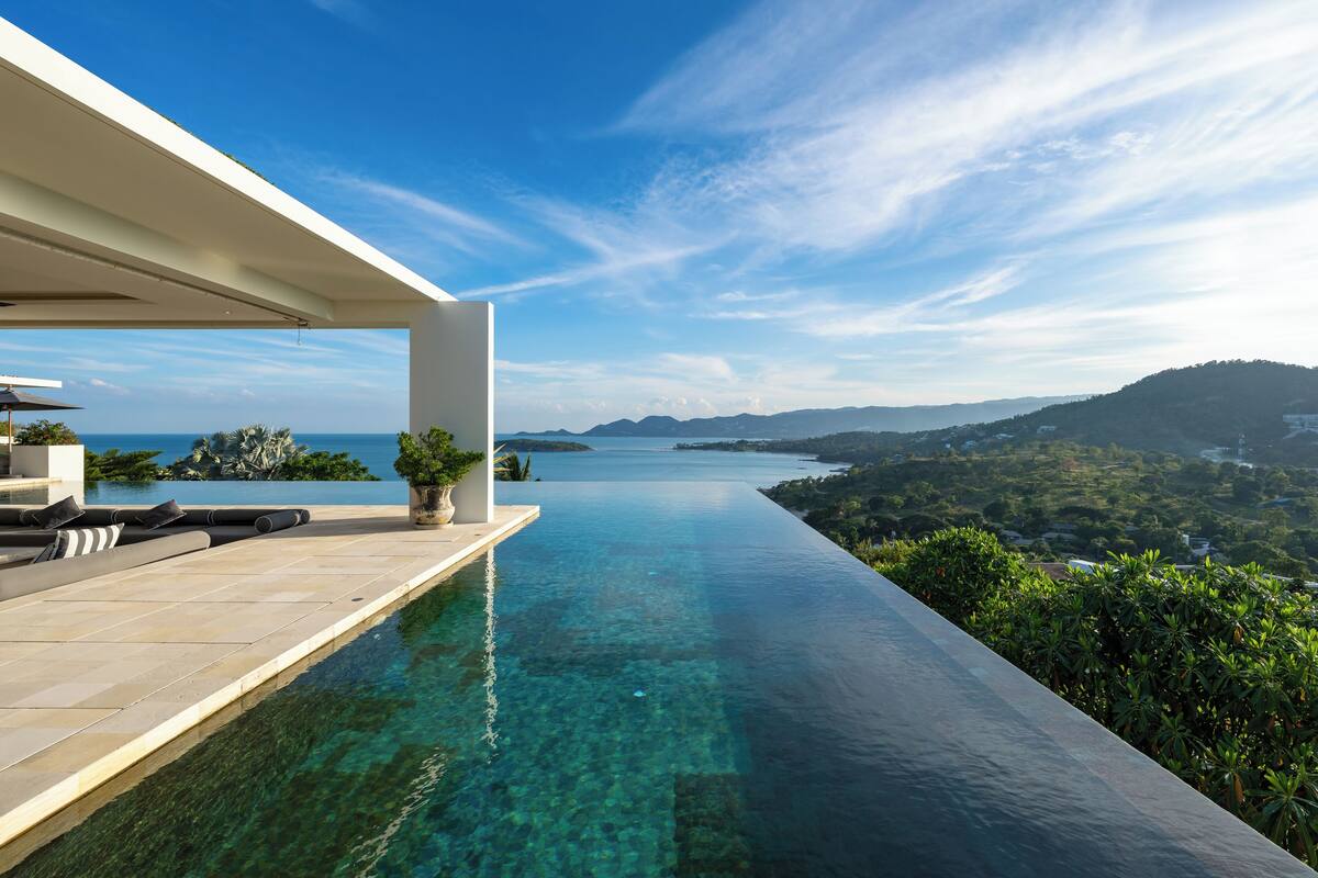 Uma piscina infinita moderna com água cristalina, uma estrutura branca de um lado e vista para a costa distante e colinas.