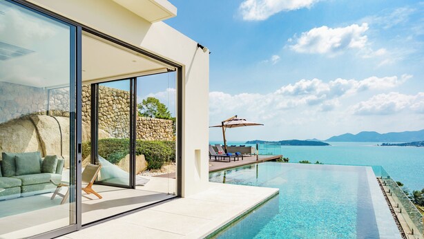 A modern infinity pool with a clear view of the sea and mountains, a glass-enclosed seating area, and a stone wall.