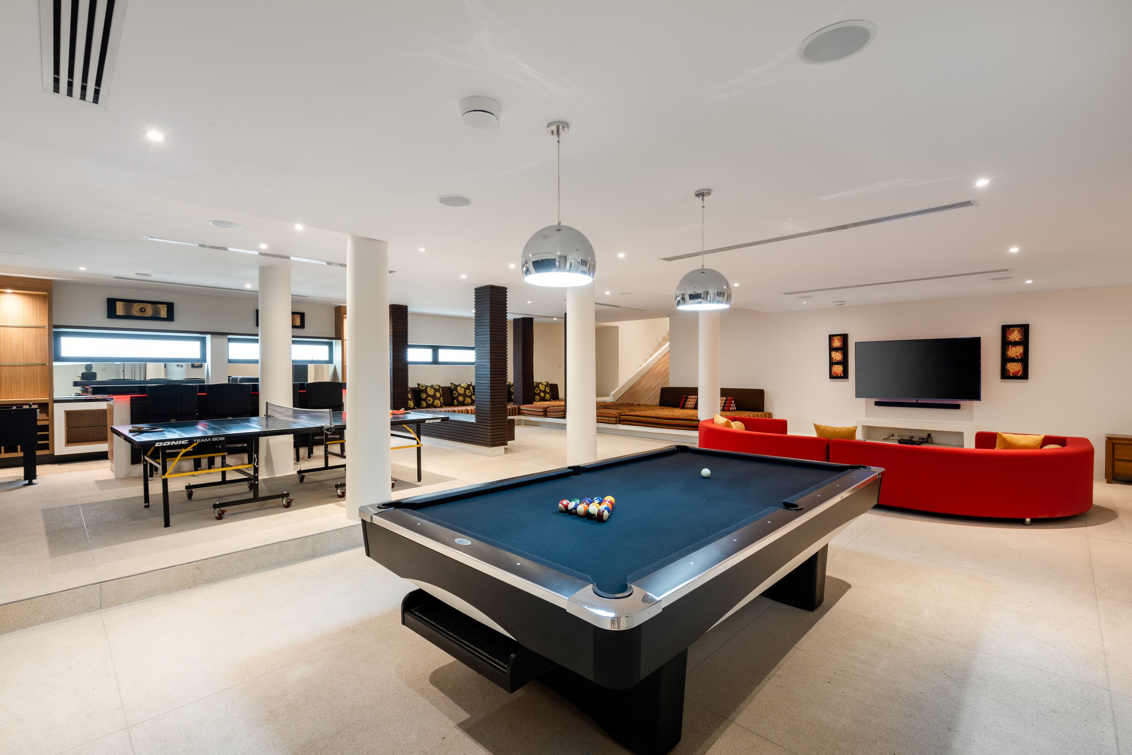 A modern indoor pool table with a billiard cue and balls, a red sofa, and a flat-screen TV mounted on the wall.