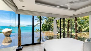 A bedroom with a large bed, a ceiling fan, and a view of the ocean and palm trees through floor-to-ceiling windows.