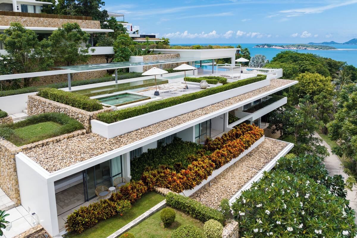 A modern multi-story building with a rooftop pool, lush landscaping, and a view of the ocean.