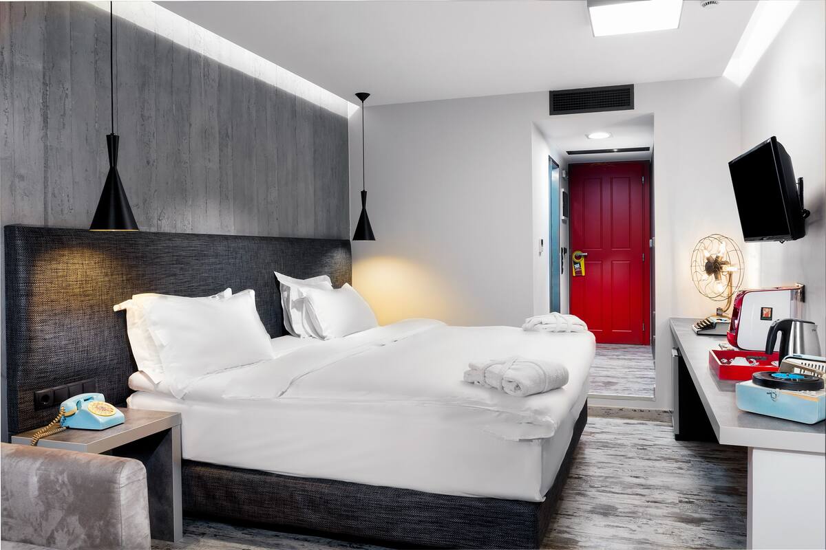 A modern hotel room with a large bed, bedside table, and a desk with a flat-screen TV.