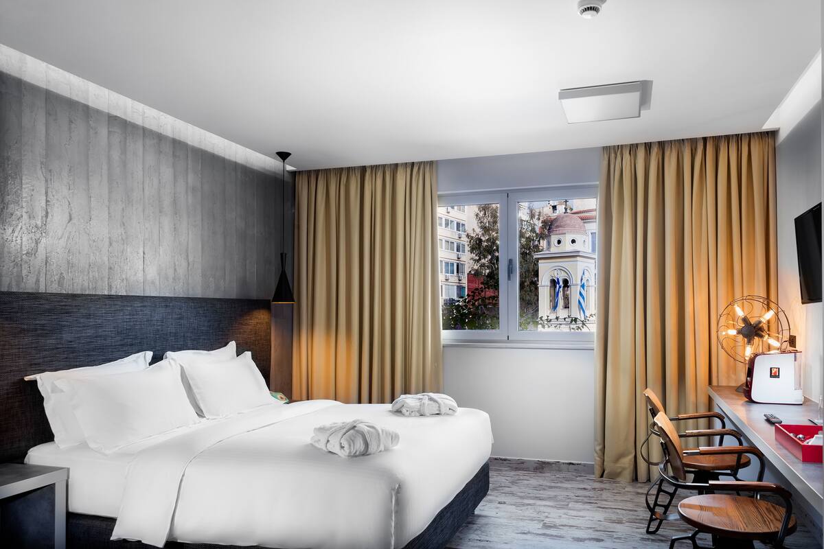 A modern hotel room with a large bed, a desk with a chair, a window with curtains, and a view of buildings outside.