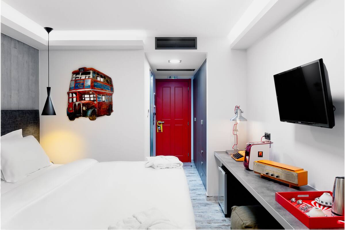 A hotel room with a bed, a red door, a flat-screen TV, a desk with a lamp, and a vintage radio.