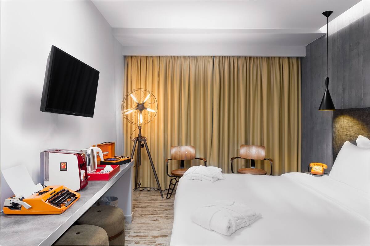 A hotel room with a bed, a desk with a typewriter, a desk lamp, a fan, a telephone, and a TV mounted on the wall.