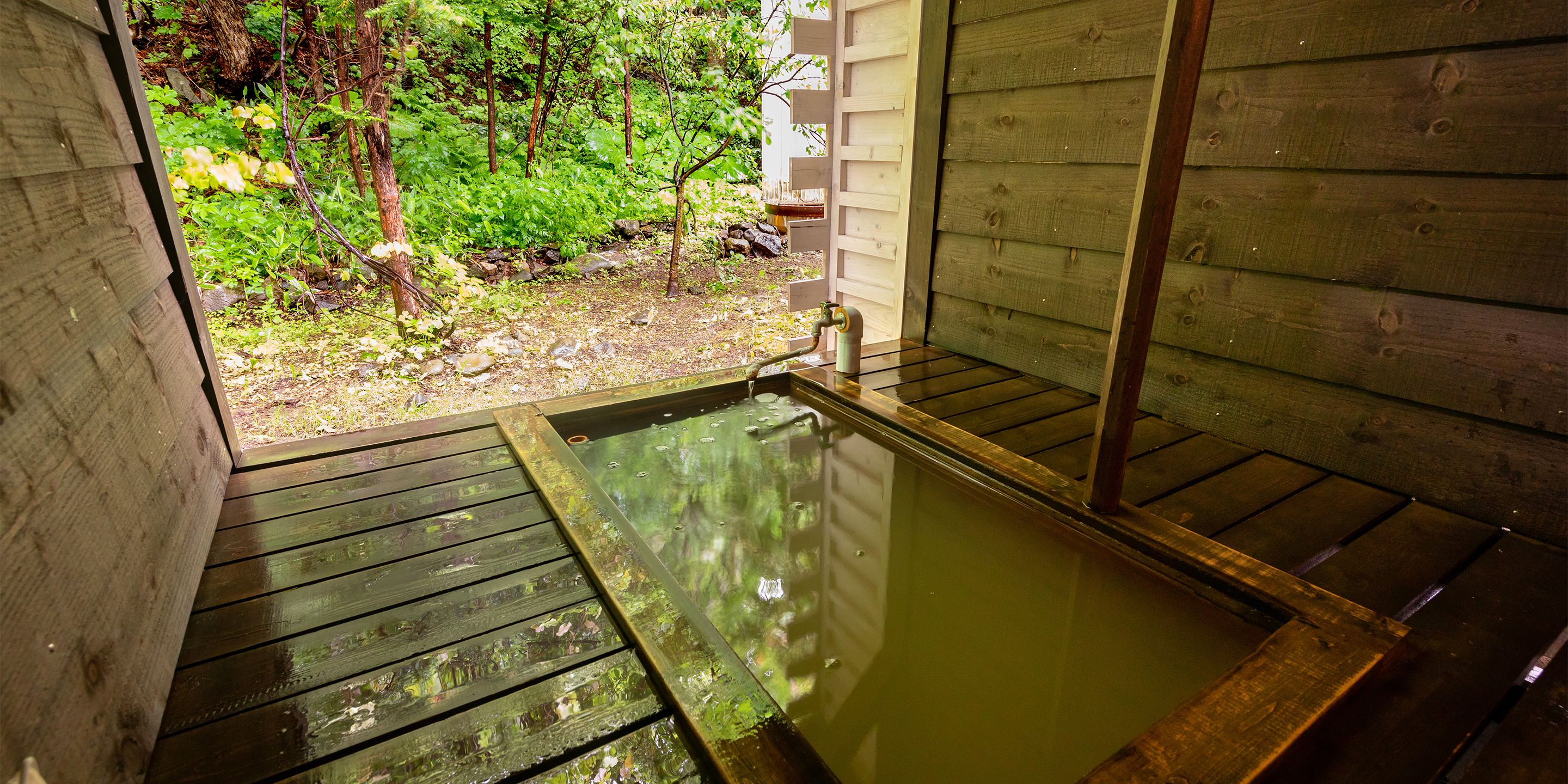 Une terrasse en bois mène à un espace de baignade dans une source d’eau chaude, avec une clôture en bois et de la verdure en arrière-plan.
