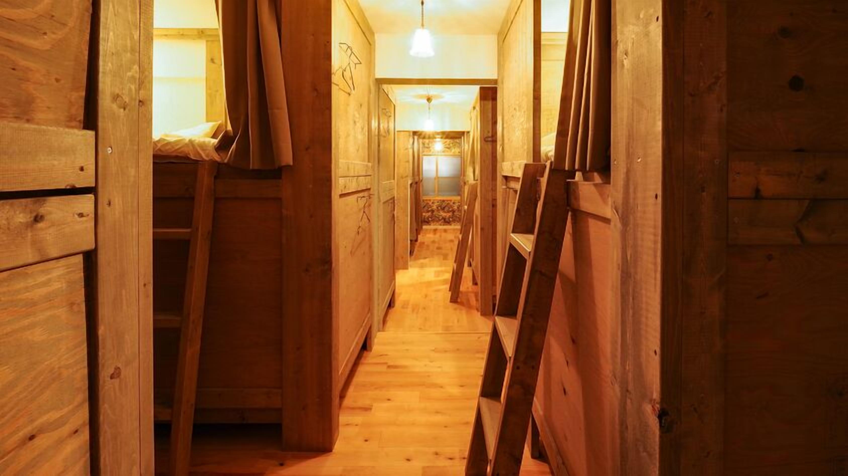 Ein schmaler Holzgang mit Stockbetten und einer Leiter, der zu einem Zimmer mit einem Bett und einer Pendelleuchte führt.