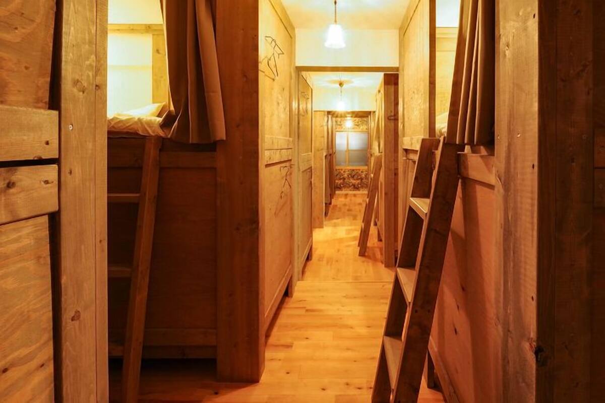 A narrow wooden corridor with bunk beds and a ladder, leading to a room with a bed and a hanging light fixture.