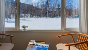 A room with a large window offering a view of a snowy landscape with trees and a cabin. There are two wooden chairs and a small table with a cup of coffee and a brochure. - Shiretoko Village (Shari)
