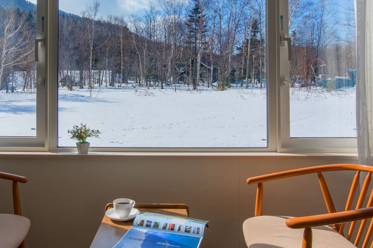 A room with a large window offering a view of a snowy landscape with trees and a cabin. There are two wooden chairs and a small table with a cup of coffee and a brochure.