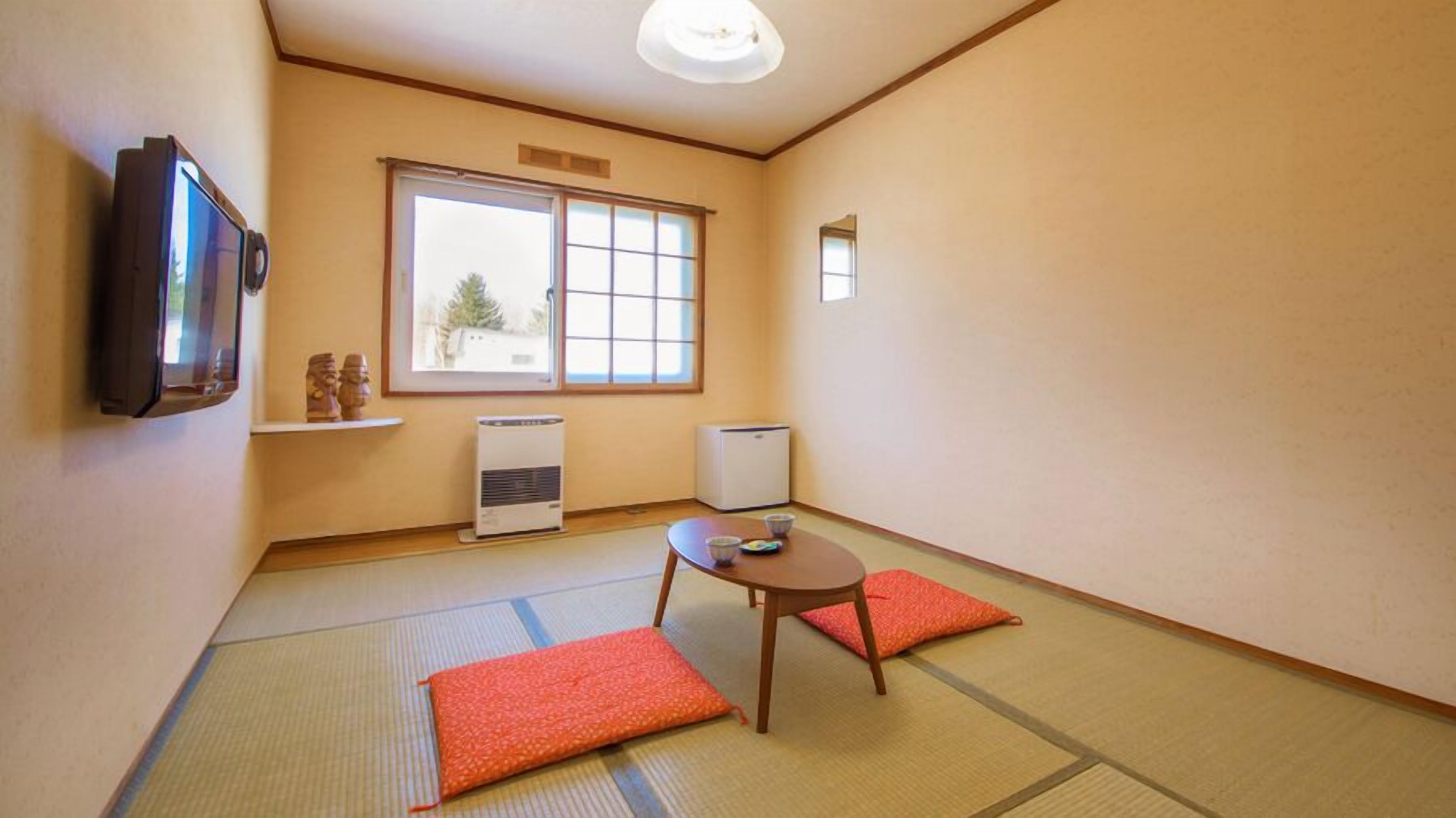 Ein Zimmer mit Tatami-Boden, einem Fernseher, einem Heizgerät, einem kleinen Tisch mit Tassen und einem Fenster mit Aussicht.