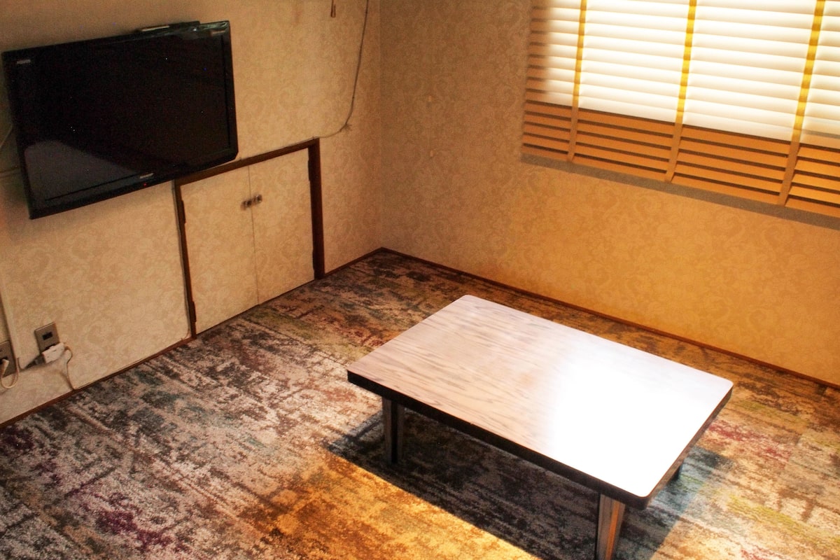 A small room with a television mounted on the wall, a wooden table, and a patterned carpet.