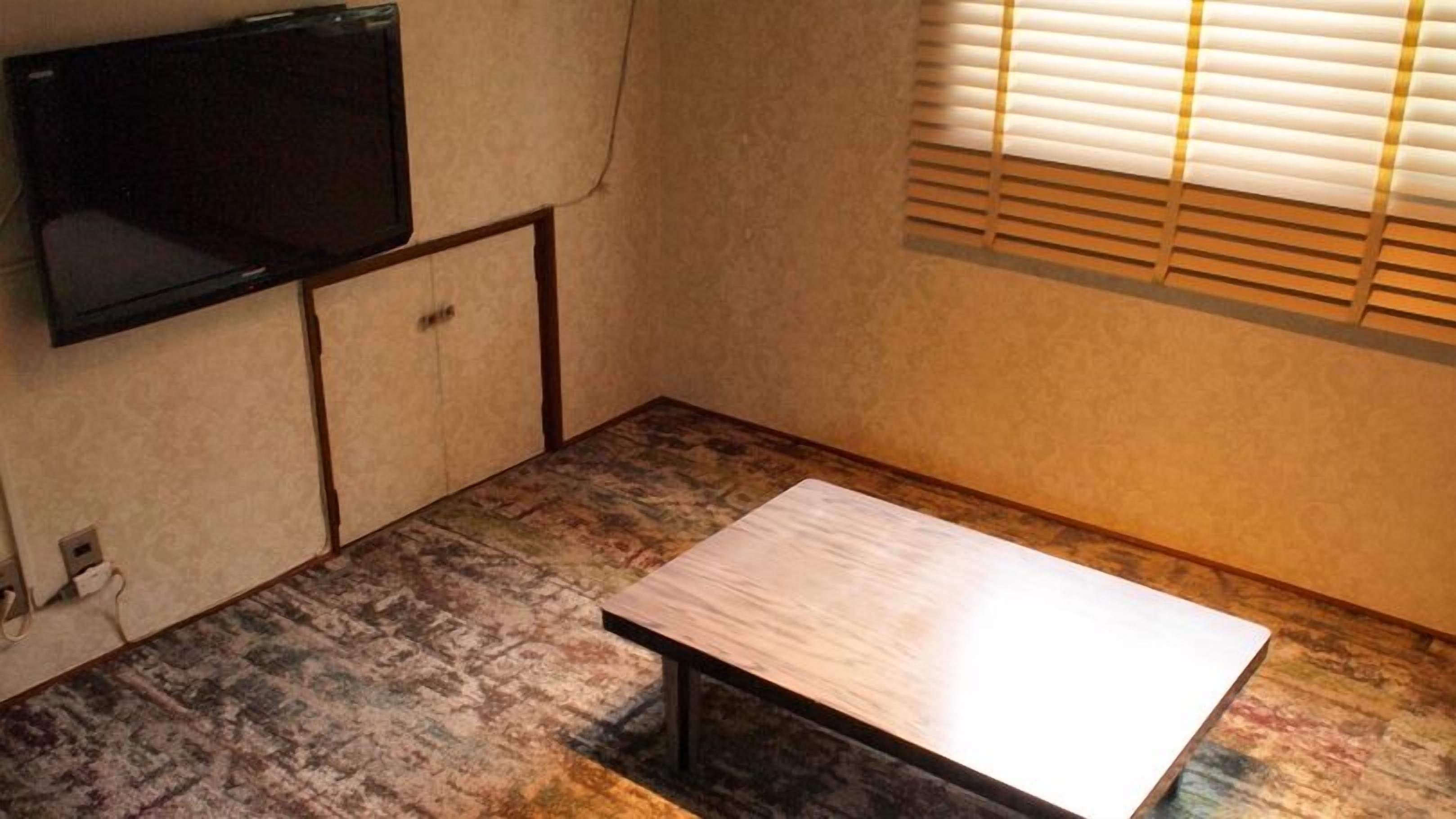 Ein kleines Zimmer mit Fernseher an der Wand, einem Holztisch und einem Fenster mit Jalousien.