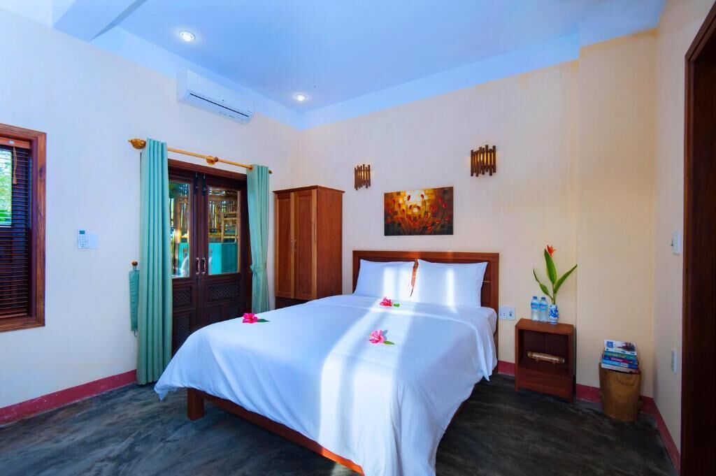 A neatly arranged hotel room with a large bed, a wooden wardrobe, a small bedside table with a vase, and a door with a green curtain.